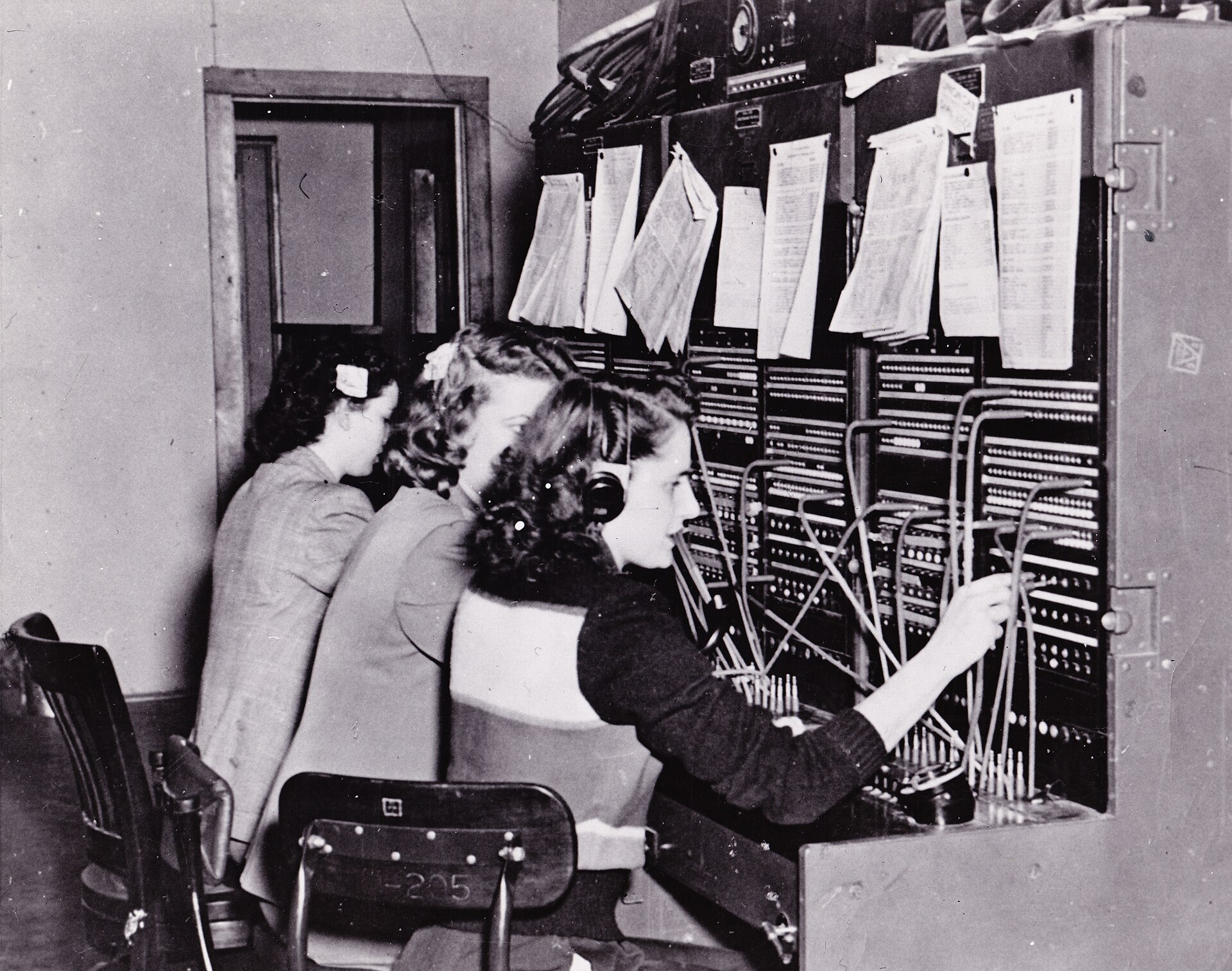 Call records clipped in sheaves along the top of the Fort Richardson switchboard in 1950, operators connecting calls below.